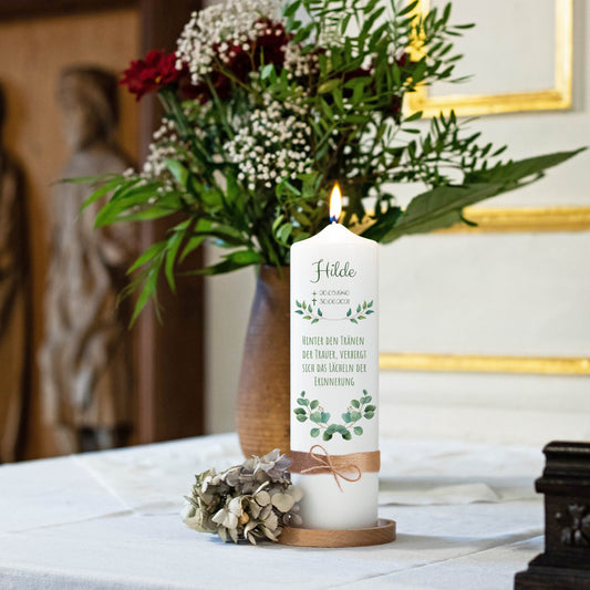 Brennende Trauerkerze mit Name, Lebensdaten und Spruch auf einem Tisch mit Blumen