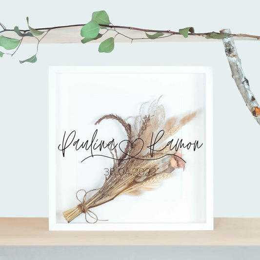 Weißer Brautstraußrahmen mit Namen in feiner Handschrift, kleinem Herz und Datum über Trockenblumen