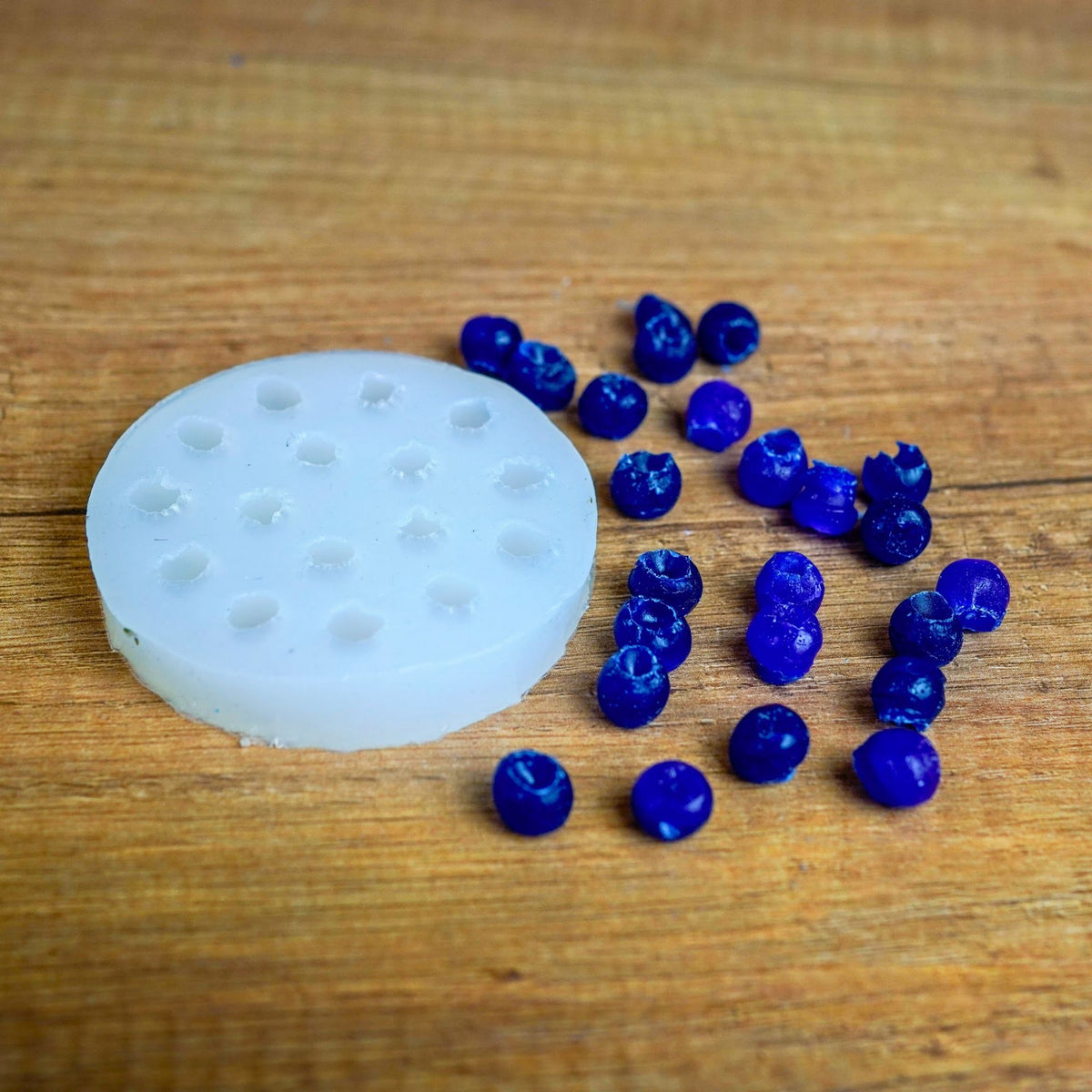 Runde weiße Silikonform mit Mini-Beeren-Mulden, daneben viele blau gegossene Wachsbeeren auf Holz
