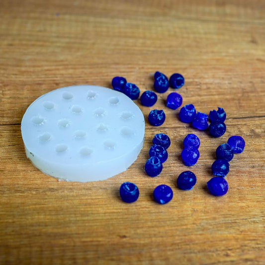 Runde weiße Silikonform mit Mini-Beeren-Mulden, daneben viele blau gegossene Wachsbeeren auf Holz