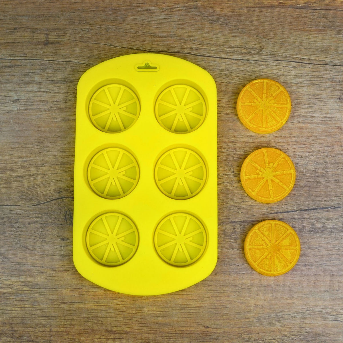 Gelbe Silikonform mit 6 Zitrusscheiben-Mulden, daneben drei fertig gegossene orange Fruchtscheiben